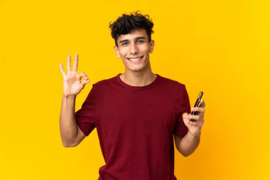 Arjantinli genç adam sarı arka planda cep telefonu kullanıyor ve tamam işareti yapıyor.