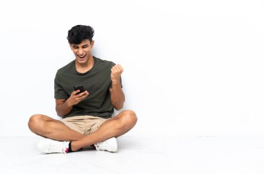 Arjantinli genç adam zafer pozisyonunda telefonla yerde oturuyor.
