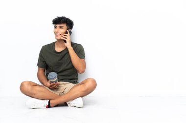 Genç Arjantinli bir adam elinde kahve ve cep telefonu ile yerde oturuyor.