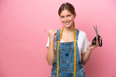 Pembe arka planda izole edilmiş genç beyaz kadın bir ürünü sunmak için kenarı işaret ediyor.