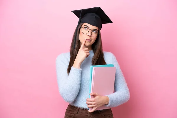 年轻的巴西女学生戴着一顶带粉色背景的毕业帽 抬起头来时感到疑惑 — 图库照片
