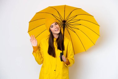 Yağmura dayanıklı ceketi ve şemsiyesi olan genç Brezilyalı kadın beyaz arka planda izole edilmiş mutlu bir ifadeyle selam veriyor.