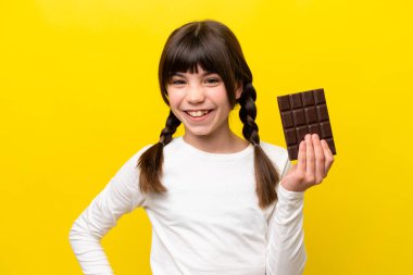 Sarı arka planda izole edilmiş küçük beyaz kız çikolata tableti alıyor ve mutlu.