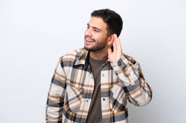 Genç Brezilyalı bir adam beyaz arka planda izole edilmiş bir şekilde elini kulağına koyarak bir şeyler dinliyor.