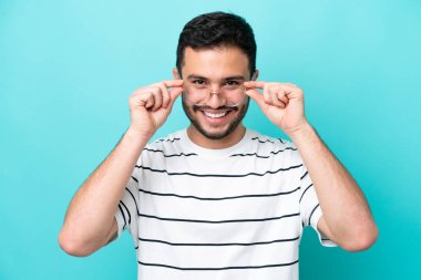 Genç Brezilyalı bir adam mavi arka planda izole edilmiş, mutlu bir ifadeyle gözlüklü.