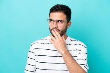 Genç Brezilyalı bir adam mavi arka planda gözlüklü ve kuşkulu.