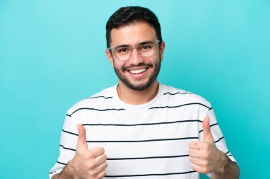 Mavi arka planda gözlüklü ve başparmağıyla izole edilmiş genç Brezilyalı bir adam.