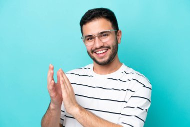 Mavi arka planda gözlüklü ve alkışlayan genç Brezilyalı adam.