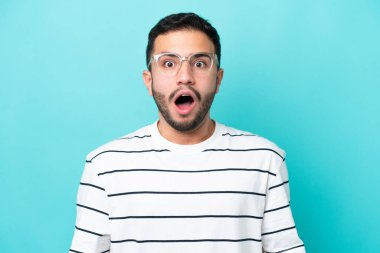 Genç Brezilyalı bir adam mavi arka planda izole edilmiş gözlüklü ve şaşırmış bir ifadeyle