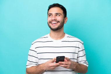 Genç Brezilyalı bir adam mavi arka planda cep telefonu kullanarak izole edilmiş.