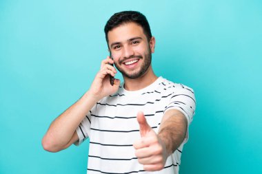 Genç Brezilyalı adam mavi arka planda izole edilmiş. Başparmaklarını kaldırırken cep telefonuyla sohbet ediyor.