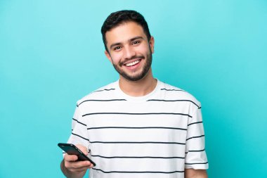Genç Brezilyalı adam mavi arka planda cep telefonu kullanarak izole edilmiş.