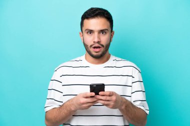 Genç Brezilyalı adam mavi arka planda izole edilmiş kameraya bakarken şaşırmış bir ifadeyle cep telefonuna bakıyor.