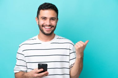 Genç Brezilyalı adam mavi arka planda izole edilmiş cep telefonu kullanıyor ve yan tarafı işaret ediyor.