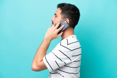 Genç Brezilyalı bir adam mavi arka planda izole edilmiş. Cep telefonuyla biriyle sohbet ediyor.