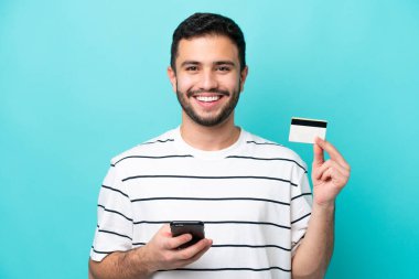 Genç Brezilyalı bir adam mavi arka planda izole edilmiş cep telefonuyla kredi kartı satın alıyor.