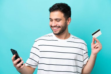 Genç Brezilyalı bir adam mavi arka planda izole edilmiş cep telefonuyla kredi kartı satın alıyor.