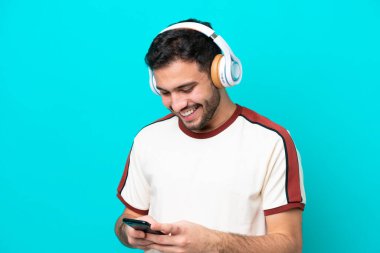 Genç Brezilyalı adam mavi arka planda izole edilmiş müzik dinliyor ve cep telefonu arıyor.
