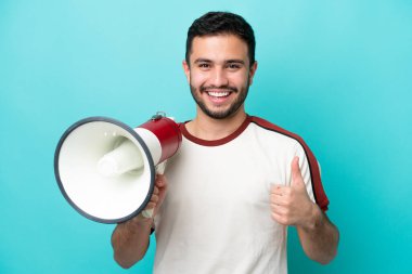 Mavi arka planda izole edilmiş genç Brezilyalı bir adam baş parmağıyla bir megafon tutuyor.