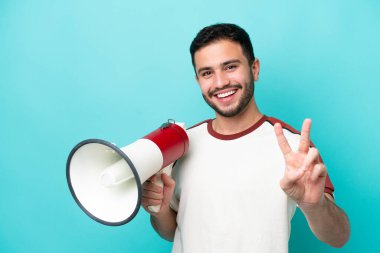Genç Brezilyalı bir adam mavi arka planda izole edilmiş elinde bir megafonla gülümsüyor ve zafer işareti gösteriyor.