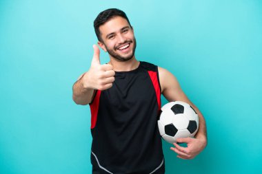 Genç futbolcu Brezilyalı adam mavi arka planda izole edildi. Başparmakları havada çünkü iyi bir şey oldu.