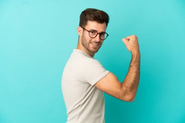 Mavi arka planda izole edilmiş beyaz, yakışıklı genç bir adam güçlü bir jest yapıyor.