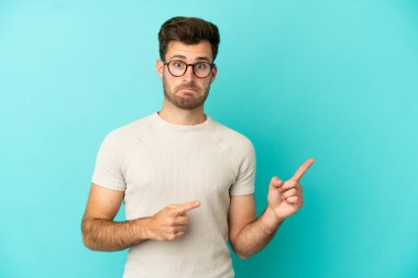 Kafkasyalı yakışıklı adam mavi arka planda izole edilmiş, korkmuş ve kenarı işaret ediyor.