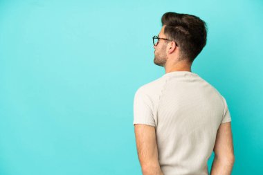 Kafkasyalı yakışıklı genç adam mavi arka planda izole edilmiş ve arkasına bakıyor.