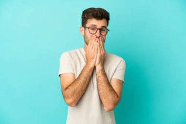 Genç, beyaz, yakışıklı bir adam mavi arka planda izole edilmiş mutlu ve gülümseyen elleriyle ağzını kapatıyor.