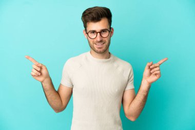Mavi arka planda izole edilmiş beyaz tenli yakışıklı adam parmak uçlarını işaret ediyor ve mutlu.