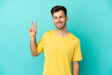 Mavi arka planda izole edilmiş genç beyaz adam gülümsüyor ve zafer işareti gösteriyor.