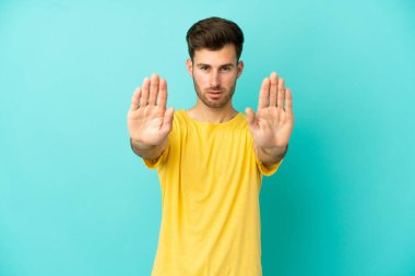 Beyaz tenli yakışıklı adam mavi arka planda izole edilmiş. Durma hareketi yapıyor ve hayal kırıklığına uğruyor.