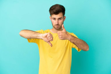 Beyaz tenli yakışıklı adam mavi arka planda izole edilmiş, iyiye-kötüye işaret yapıyor. Evet ya da hayır arasında kararsız