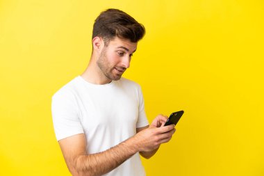 Sarı arka planda izole edilmiş genç beyaz adam cep telefonuyla mesaj ya da e-posta gönderiyor.