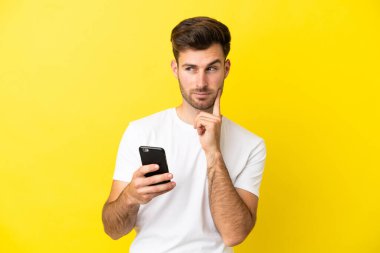 Genç, beyaz, yakışıklı bir adam sarı arka planda cep telefonu kullanıyor ve düşünüyor.