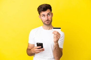 Beyaz tenli yakışıklı adam sarı arka planda izole edilmiş. Cep telefonuyla kredi kartıyla alışveriş yapıyor.