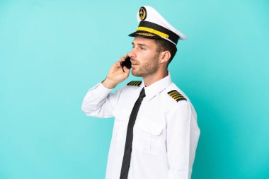 Kafkasyalı uçak pilotu mavi arka planda izole edilmiş cep telefonuyla sohbet ediyor.