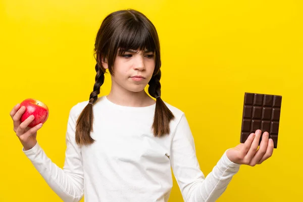 Malá Běloška Izolovaná Žlutém Pozadí Pochybnostmi Při Užívání Čokoládové Tablety — Stock fotografie