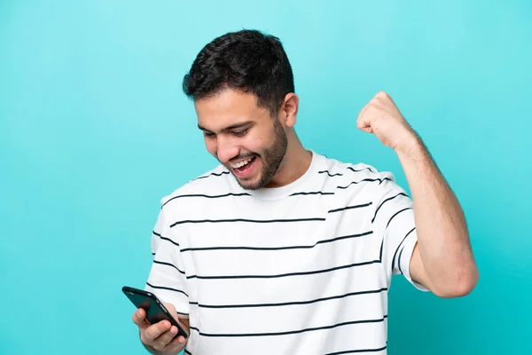 Junger Brasilianer Isoliert Auf Blauem Hintergrund Mit Handy Und Siegesgeste — Stockfoto