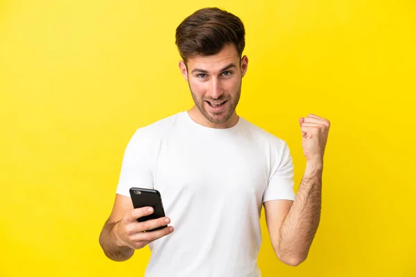 年轻的高加索帅哥在黄色背景下被隔离使用手机和做胜利的手势 — 图库照片