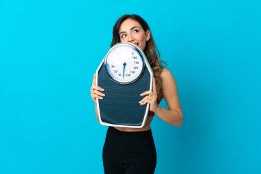 Mavi arka planda tartı tutan beyaz bir kadın. Tartma makinesi var ve arkasında saklanıyor.