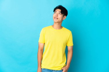 Genç Çinli bir adam mavi arka planda izole edilmiş, yukarı bakarken bir fikir düşünüyor.