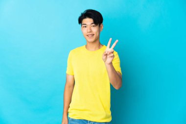 Mavi arka planda izole edilmiş genç Çinli adam gülümsüyor ve zafer işareti gösteriyor.