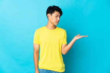 Genç Çinli adam mavi arka planda izole edilmiş. Kopyalama alanını şüpheyle tutuyor.