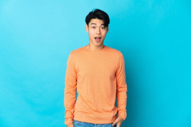 Genç Çinli bir adam mavi arka planda izole edilmiş ve yüzünde sürpriz bir ifade var.