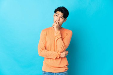 Mavi arka planda izole edilmiş genç Çinli adamın şüpheleri ve kafa karıştırıcı bir yüz ifadesi var.