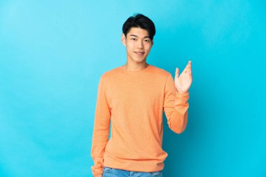 Mavi arka planda izole edilmiş genç bir Çinli, mutlu bir ifadeyle selamlıyor.