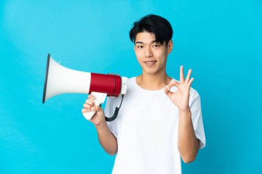 Mavi arka planda izole edilmiş genç bir Çinli elinde bir megafonla tamam işaretini parmaklarıyla gösteriyor.