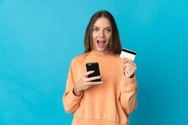 Genç Litvanyalı bir kadın mavi arka planda izole edilmiş cep telefonuyla alışveriş yapıyor ve elinde şaşırmış bir ifadeyle bir kredi kartı tutuyor.
