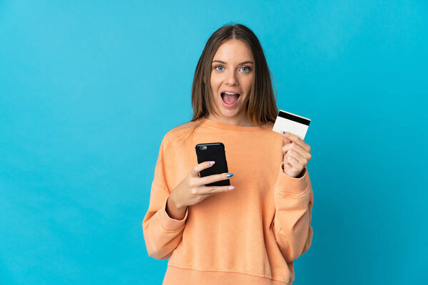 Young Lithuanian woman isolated on blue background buying with the mobile and holding a credit card with surprised expression
